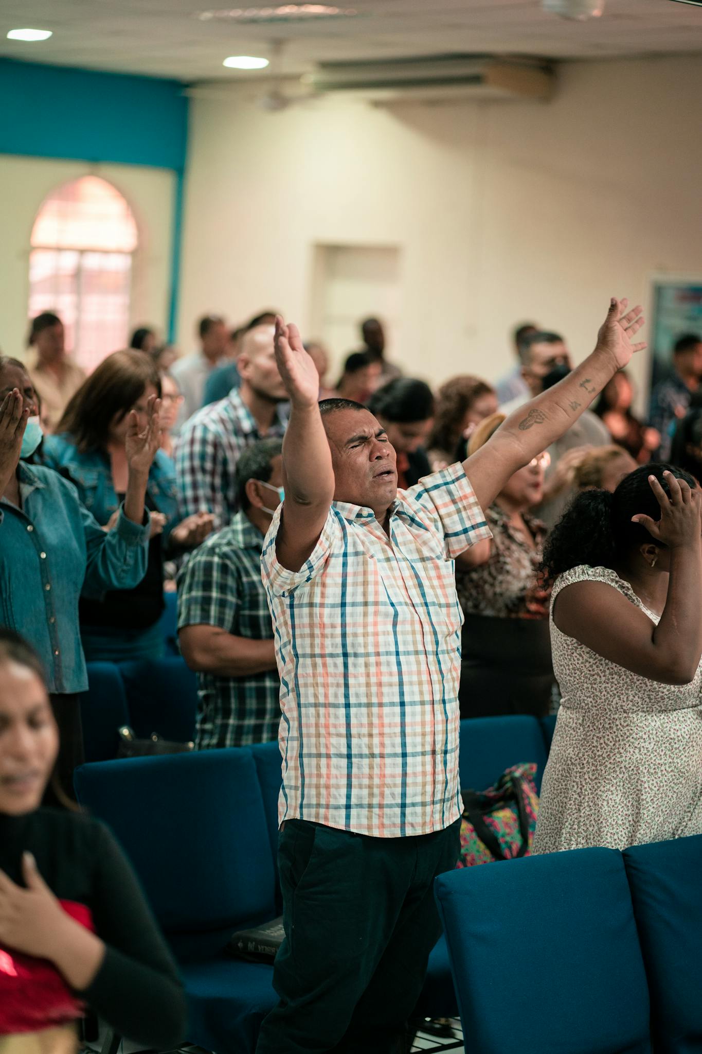 A diverse group of people engaged in prayer indoors, expressing emotions with arms raised.